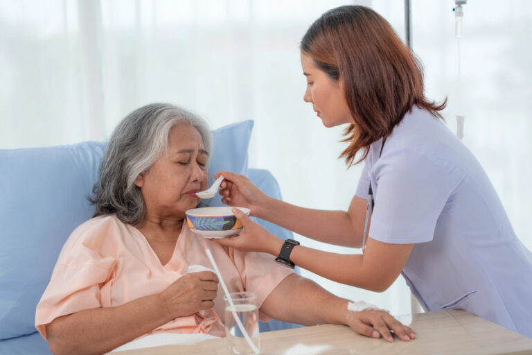 Nurse feeding an elderly woman with a spoon in a hospital room. The woman is wearing a pink gown and sitting in bed. A glass of water is on the table nearby.