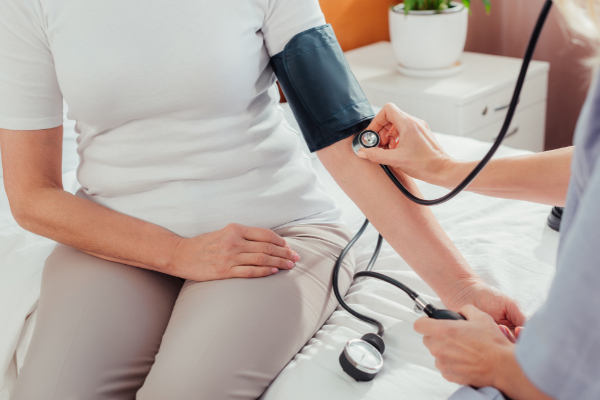 A person is seated on a bed having their blood pressure checked with a cuff and stethoscope.