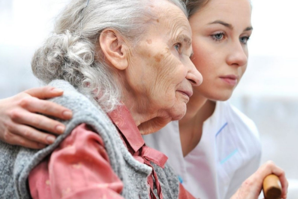 Elderly woman with gray hair and a red shirt supported by a young woman in a white top, both looking attentively in the same direction.