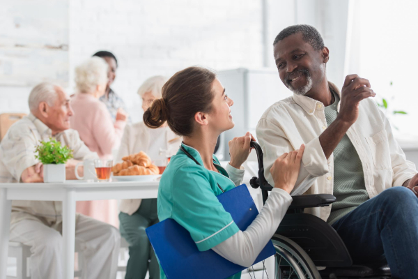 A caregiver in a green uniform talks to a man in a wheelchair, smiling. In the background, a group of elderly people sits at a table with food and drinks.