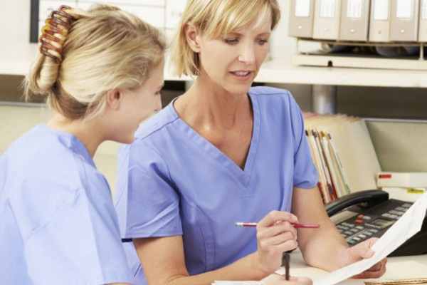 Two healthcare workers in blue scrubs reviewing documents at a desk in a medical office.