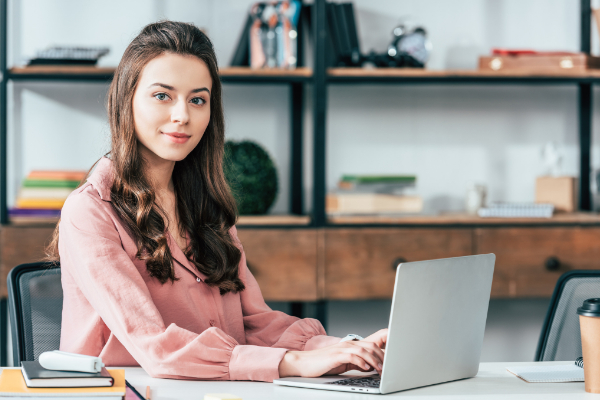 A woman in a pink blouse sits at a desk, using a laptop. Shelves with books and decorative items are in the background.