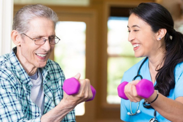 An elderly man and a nurse smile as they lift purple dumbbells together in a bright room.