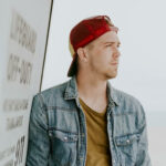 A man wearing a red cap and denim jacket leans against a wall with a lifeguard sign. He gazes into the distance.