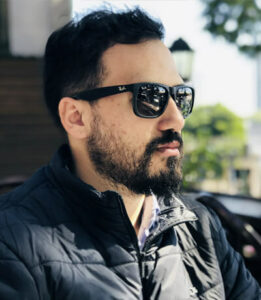 Man with dark hair and beard wearing sunglasses and a black jacket, sitting outdoors.