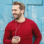 Man in a red sweater smiling while holding a smartphone and wearing earbuds, standing against a blue brick wall.