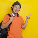 Person in an orange shirt and beanie smiles while pointing to the right. They have headphones around their neck and a backpack on one shoulder. Yellow background.