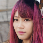 A person with long, bright pink hair and a slight smile looks towards the camera. The background appears blurred.