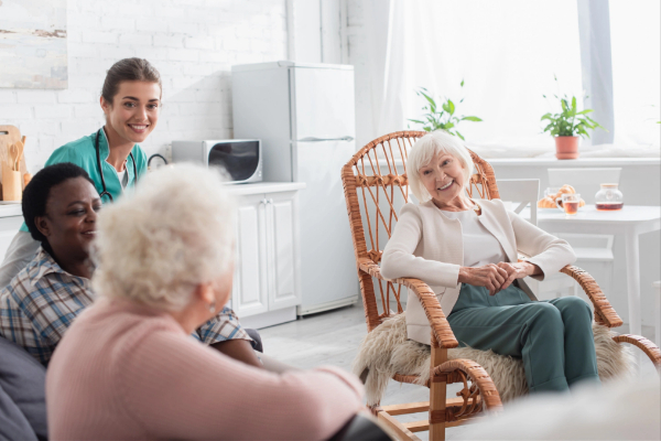 A group of elderly people sit and chat in a bright, cozy room with a young caregiver standing nearby, smiling.
