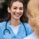 A smiling person wearing a blue medical uniform and stethoscope, standing outdoors and talking to someone.