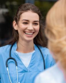 A smiling person wearing a blue medical uniform and stethoscope, standing outdoors and talking to someone.