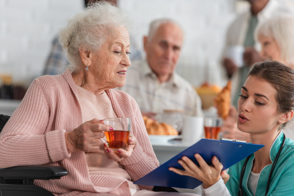 Elderly woman in a wheelchair holds a cup of tea while a young caregiver in scrubs shows her a document on a clipboard. Other people are visible in the background.