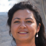 A woman with long dark hair and pearl earrings smiles outdoors with a blurred background.