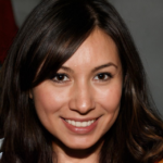 Smiling person with long brown hair and a dark top against a neutral background.