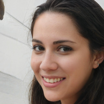 A woman with long dark hair smiles while looking at the camera. The background is a light colored wall. A woman with long dark hair smiles while looking at the camera. The background is a light colored wall.